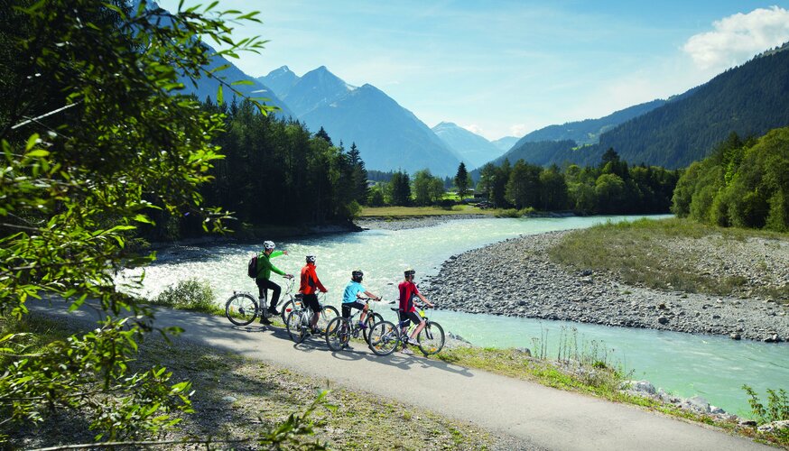 Vier Fahradfahrer:innen vor einem Fluss und Bergkulisse.