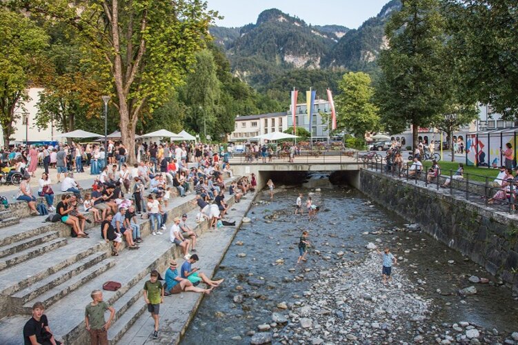 Menschen sitzen links auf Treppen und Kinder spielen im Fluss davor.