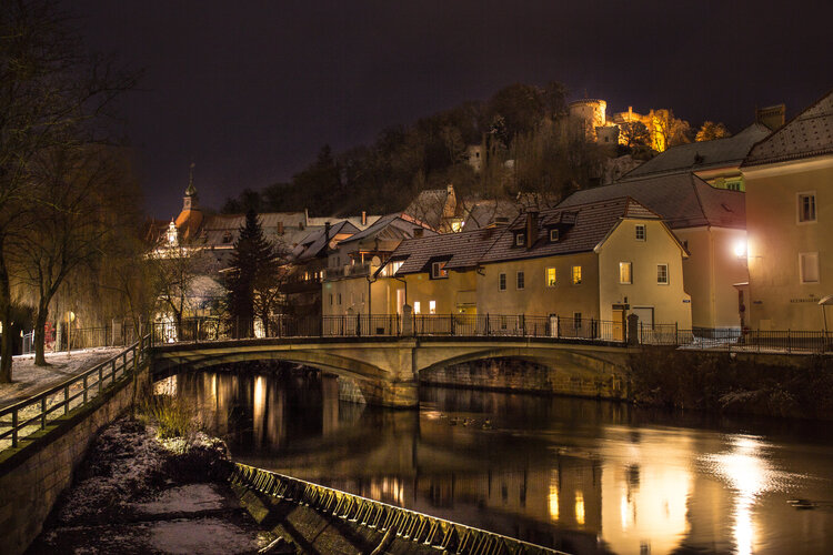 Beleuchtete Brücke und Gebäude an der Lavant in der Nacht.