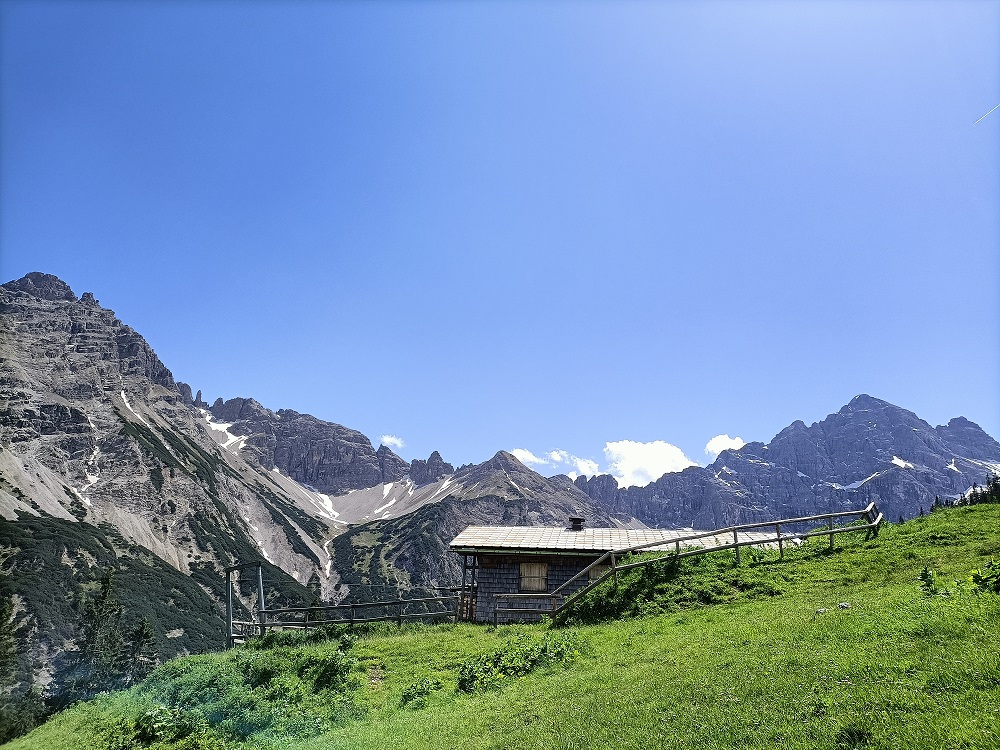 Auf einer grünen Wiese steht eine Hütte und dahinter ist eine Bergkette.